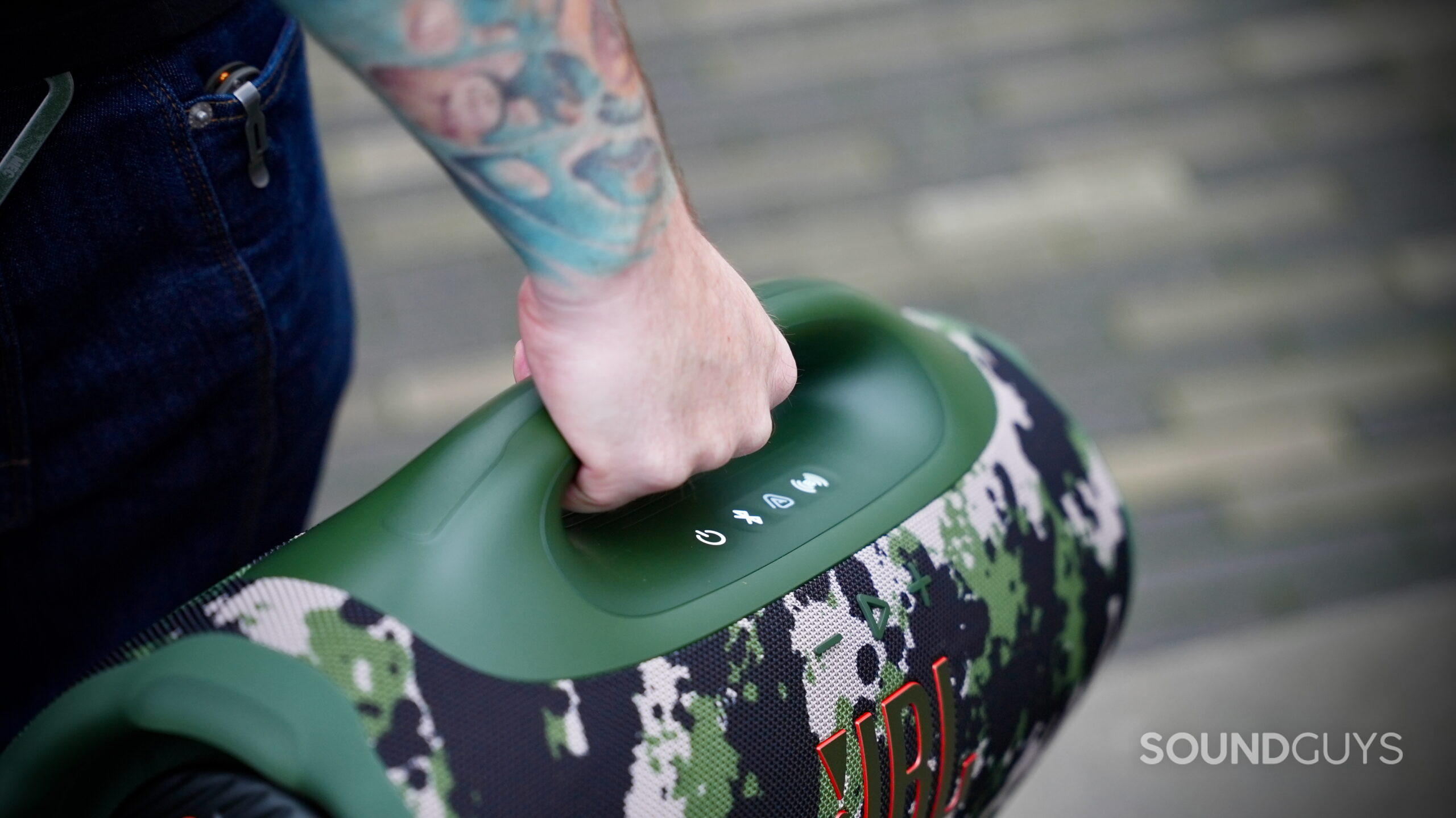 A hand carrying a camo colored JBL Boombox 4 outside